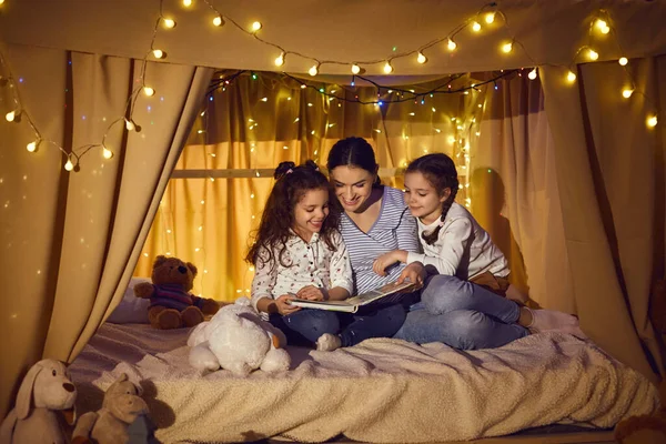 Bonne mère et les enfants assis dans la tente de la salle de jeux et le livre de lecture de contes de fées — Photo