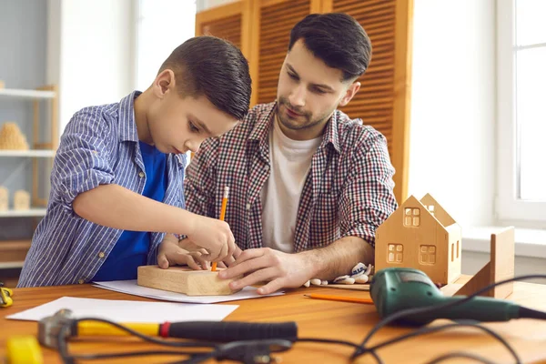 Caring father carpenter in the workshop teaches his little interested son to make a wooden house.