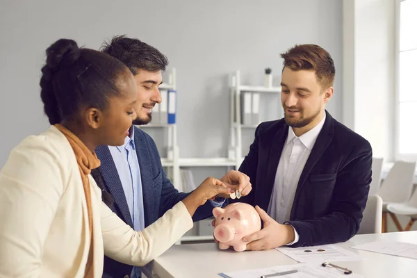 Happy couple together put money in piggy bank sitting at desk with banking advisor