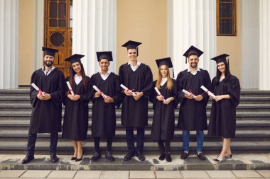 Üniversitenin arka planında diplomalarla poz veren akademik kıyafetli bir grup mezun