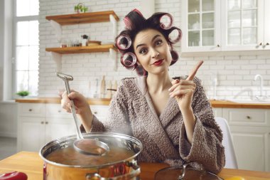 Bukleli ve bornozlu genç ve çekici ev hanımı ev mutfağında yemek yapıyor.