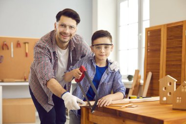 Baba ve oğul marangoz atölyesinde tahta keserken koruyucu gözlük takıyor.