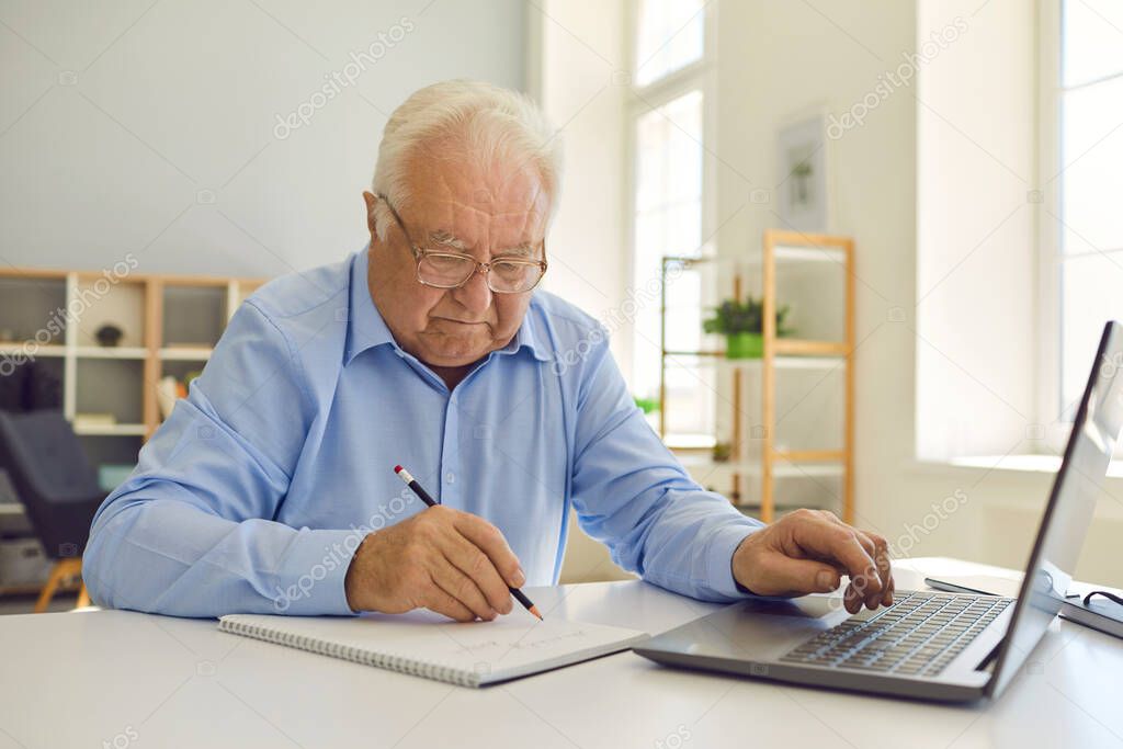Senior man in eyeglasses sitting at desk at home, using laptop and taking notes in notebook