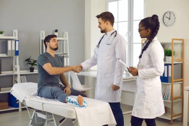 Ayak bileği incinen minnettar bir hasta doktorların elini sıkıyor ve ona yardım ettiği için teşekkür ediyor.