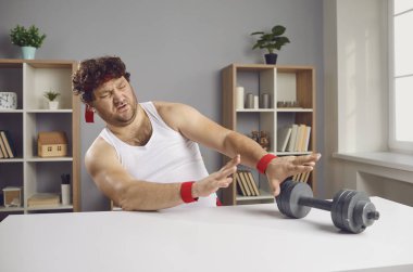 Salonda spor antrenmanlarına başlamak istemeyen komik, tembel, tombul adam dambılları itiyor.