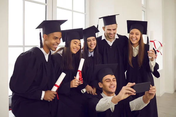 Group of happy international students with diplomas taking selfie using smartphone in classroom.