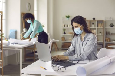 Yüz maskeli çok ırklı kadın iş arkadaşı ofiste çalışıyor. Mesafeyi koruyun.