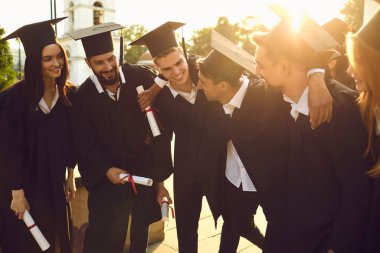 Mutlu ve başarılı üniversite mezunları akademik elbiseler içinde diplomaları ellerinde tutuyorlar ve sarılıyorlar..