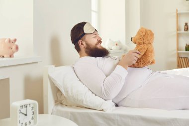 Beyaz pijamalı ve uyku maskeli neşeli bir adam uyandı ve bir çocuk gibi davranıyor..