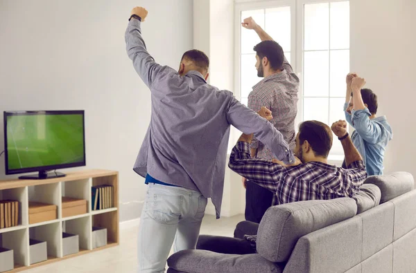 Active adult male friends with arms raised emotionally watching a football match on TV at home.
