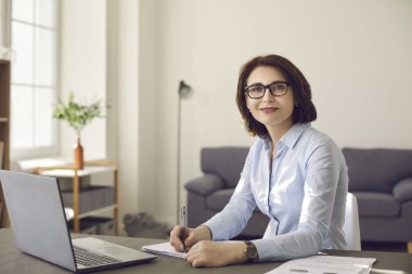 Portrait of a smiling middle aged businesswoman with glasses sitting at an office desk at work. clipart