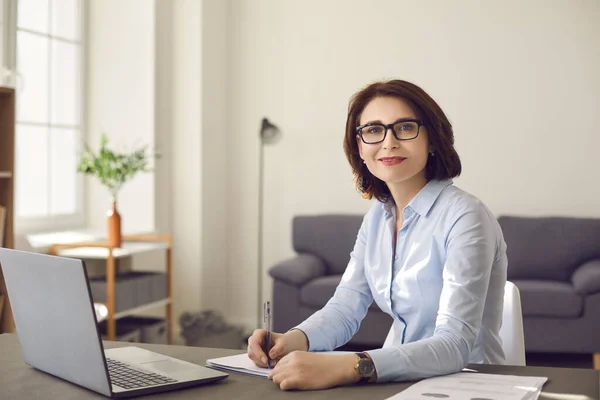 Портрет улыбающейся деловой женщины средних лет в очках, сидящей за рабочим столом. — стоковое фото