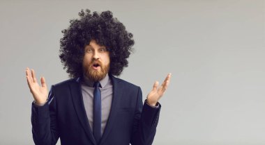 Kıvırcık peruklu komik genç adam şaşırdı. Gri fotokopi arkaplanının üzerinde duruyor.