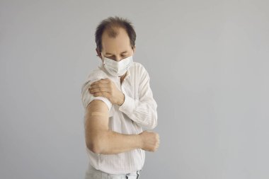 Tıbbi maske takan olgun bir adam Covid 19 aşısından sonra kolunu gösteriyor.