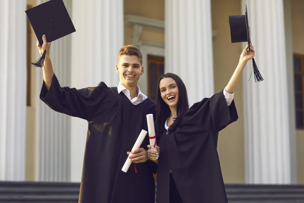 Cheerful couple girl and boy university graduates standing with dimplmas and holding bonets in raised hands