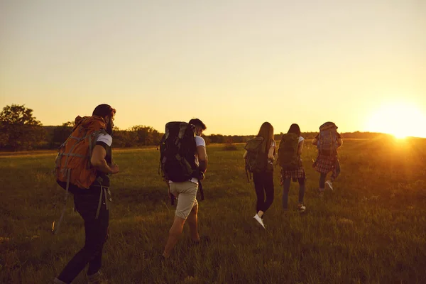 Skupina pěti lidí s velkými batohy se pohybuje v řadě přes pole při západu slunce. — Stock fotografie