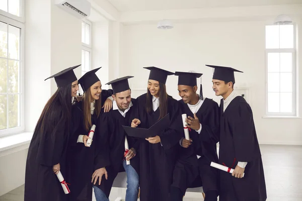 Φοιτητές διαφορετικών εθνικοτήτων ντυμένοι με φορέματα αποφοίτησης μιλούν, χαμογελούν και περιηγούνται στο άλμπουμ. — Φωτογραφία Αρχείου