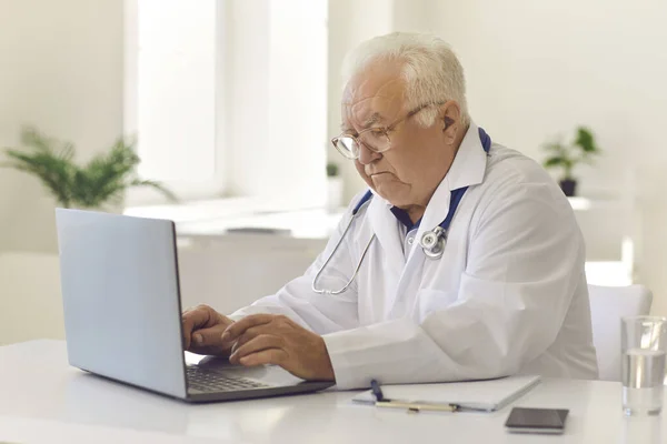 Witharige senior arts typt op laptop toetsenbord zittend aan bureau in ziekenhuis kantoor — Stockfoto