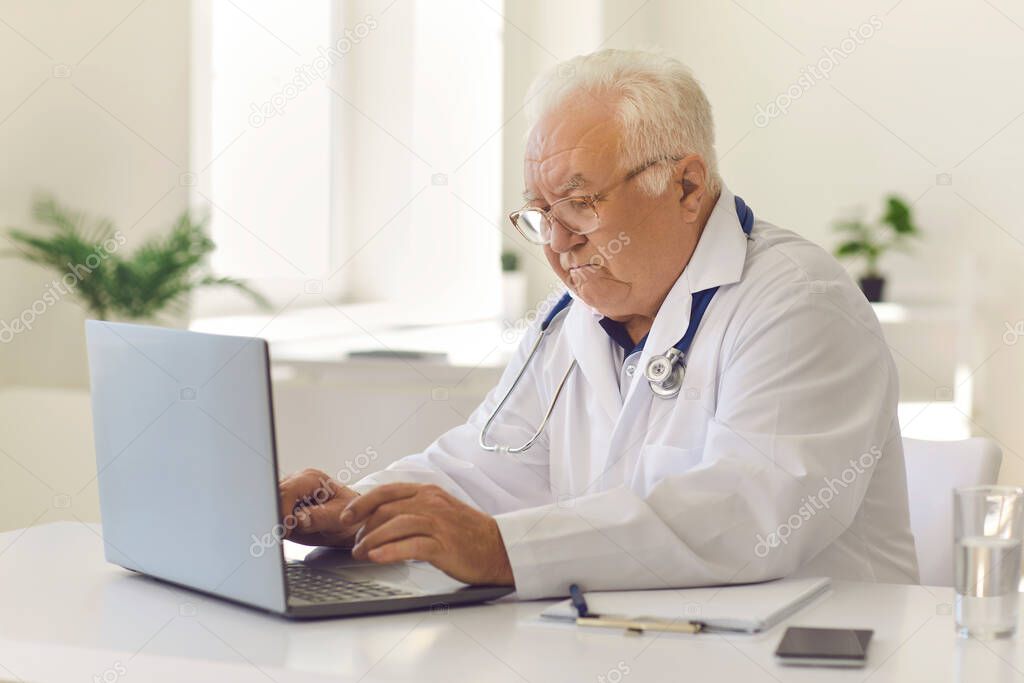 White-haired senior doctor typing on laptop keyboard sitting at desk in hospital office