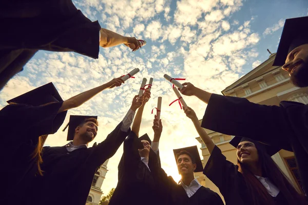 Spodní pohled absolventi zvednout své diplomy a stack je dohromady proti modré obloze s mraky. — Stock fotografie