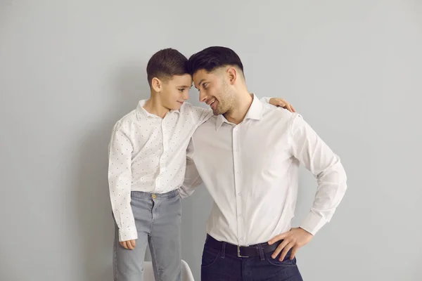 Feliz pai e filho de camisas vestido olhando um para o outro de pé no estúdio com fundo cinza — Fotografia de Stock