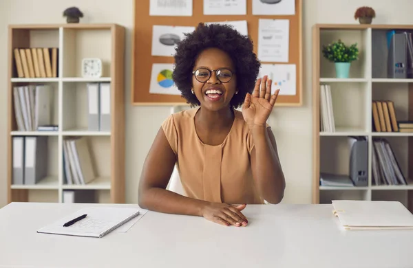 Femme afro-américaine agitant la main en regardant la caméra assise au bureau — Photo