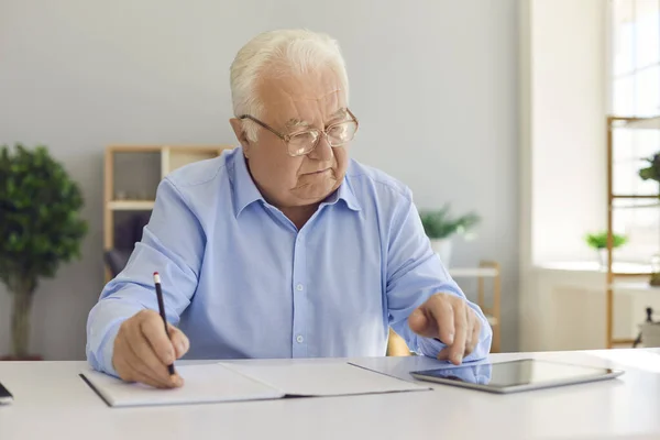 Oudere man werknemer vinden van informatie op internet op tablet en opschrijven in notebook — Stockfoto