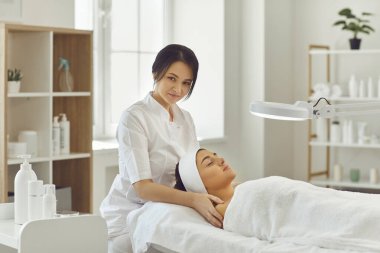 Gülümseyen doktor dermatolog genç bir kadın için rahatlatıcı yüz masajı prosedürü uyguluyor.