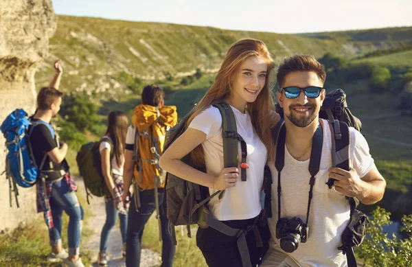 Young smiling couple hikers standing with backpacks and camera over group and rocks landscape at background — Stock fotografie