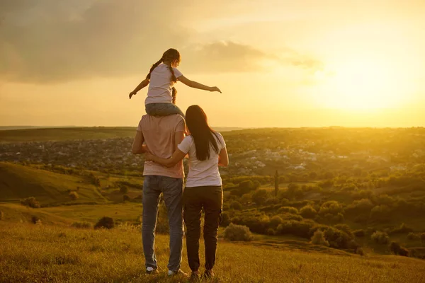 Vista traseira. Família feliz e uma criança de pé em um fundo de um pôr-do-sol à noite na natureza. — Fotografia de Stock