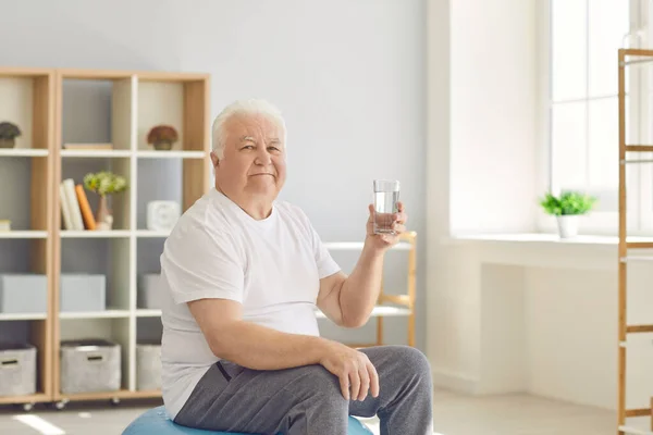 Senior man met een glas water zittend op een fitbal thuis en kijkend naar de camera. — Stockfoto