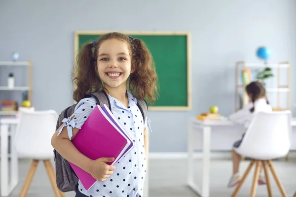 Je retourne à l'école. Écolière souriante avec un livre à la main regarde la caméra tout en étant debout en classe. — Photo