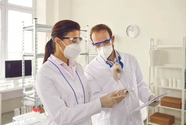 Twee wetenschappers in beschermende maskers maken notities als ze bacterieculturen in petrischaal onderzoeken — Stockfoto