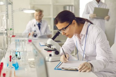 Laboratuvar önlüklü, gözlüklü ve eldivenli bilim adamı mikroskoba bakıyor ve notlar alıyor.