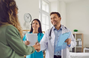 Tıbbi muayeneden sonra gülümseyen erkek doktor, genç ve mutlu hasta bir kadınla el sıkışıyor. İlaç ve sağlık hizmetleri kavramı.