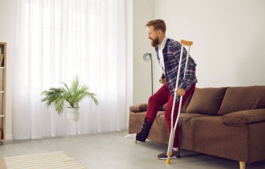 Man with leg injury leans on crutch and has difficulty getting up from couch at home. Side view of adult man with broken limb wearing bandage on his leg and having difficulty walking.