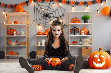 Portrait of child in spooky costume. Kid dressed up for Halloween party at home. Girl wearing black dress, witch hat and scary makeup sitting on floor in room decorated with pumpkins and spider webs