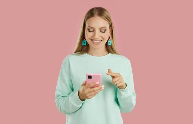 Portrait of smiling young woman using mobile phone. Cheerful blonde girl wearing sweatshirt standing and texting on smart phone, uses mobile application, shopping online over pink background
