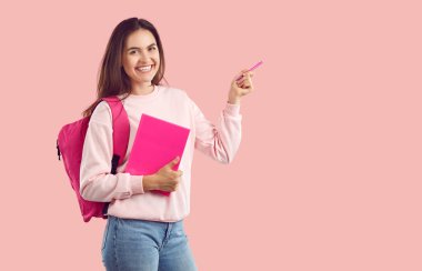 Cheerful joyful positive excited brunette college girl with bag and textbook on empty pink background points aside at homework, new course, good event, product offer, idea, exam reading recommendation