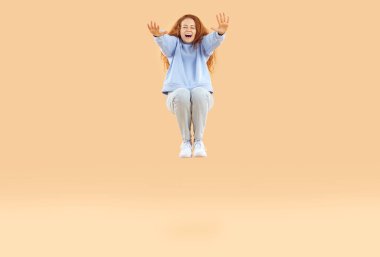 Beautiful happy red-haired young woman in light blue sweatshirt and gray jeans, squats in jump and stretches her arms forward. Studio photo of pretty girl with long wavy hair on isolated background.