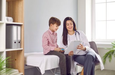 Portrait of a friendly smiling female child doctor pediatrician showing report file with appointment to a little boy patient sitting on the couch during medical examination in clinic.