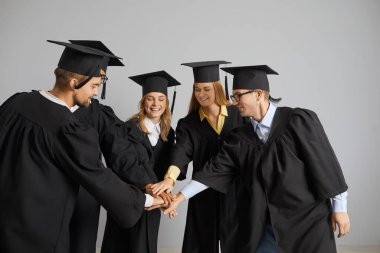 Team of happy university students and friends having fun at their graduation. Group of cheerful, joyful graduates in academic square hats and robes standing in circle and putting their hands together