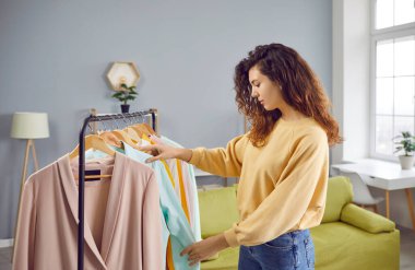 Pretty young woman choosing clothes at wardrobe at home. Attractive girl with curly long hair standing at hanger rack thinking what to wear. Fashion, clothing, style concept