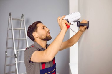 Service worker installing a modern security camera inside the house or apartment. Young man in a uniform sets up a new CCTV surveillance video camera on the wall at home