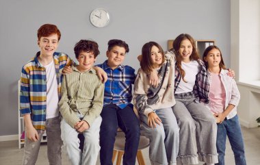 Group portrait of happy school classmates and friends in the classroom. Several children sitting on a table at school. Six happy kids sitting on a desk, hugging, looking at the camera, and smiling
