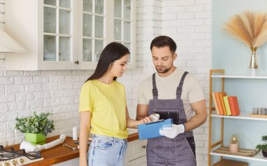 Houswife and male worker standing in kitchen wearing overall discussing, giving professional service for home repair, maintenance, plumbing change planning, focused and engaged in conversation 