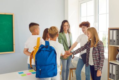 Group of school students with their young friendly female teacher standing in a circle in the classroom and holding their hands in stack together. Back to school, education and teamwork concept.