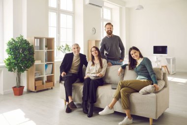 Portrait of friendly different business colleagues sitting on sofa in modern bright office. Smiling men and women of all ages are smiling while looking at camera. Concept of partnership and friendship