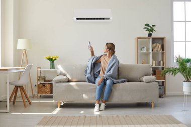 Woman whos sitting on sofa under warm plaid in living room switches off her air conditioner on wall. Young girl adjusting modern AC system, regulating temperature and enjoying cool fresh air at home
