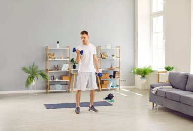 Ordinary man in morning in his home performs sports training and pumps his arm muscles. Young Caucasian man is standing in living room and using small dumbbells for sports workout. Home sports concept